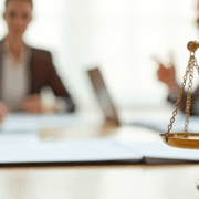 A close-up image of a brass balance scale placed on a conference table, with four blurred business professionals in suits sitting in discussion in the background. The warm lighting and shallow depth of field emphasize the symbolic scale of justice.