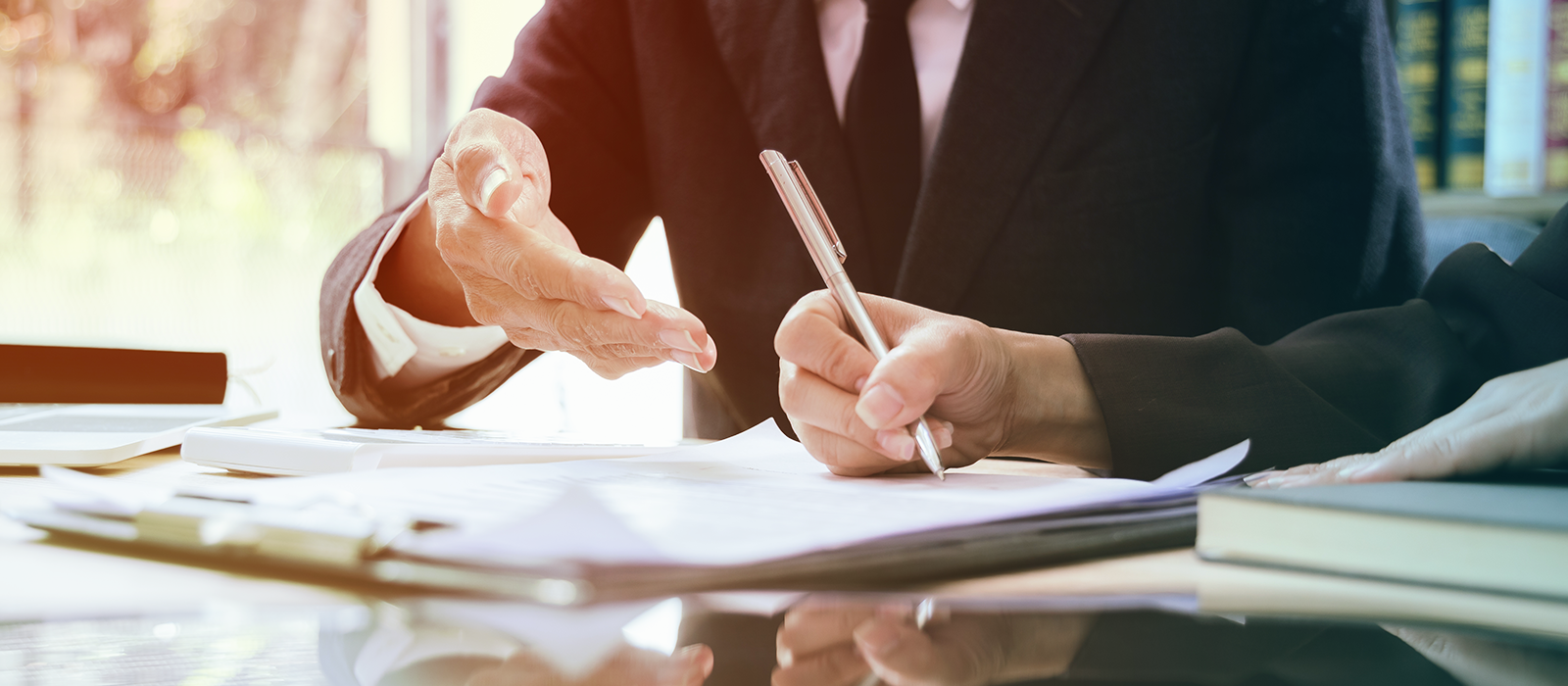 Close-up of two people in business attire reviewing and signing documents at a desk, with one gesturing while the other writes, and legal books blurred in the background.