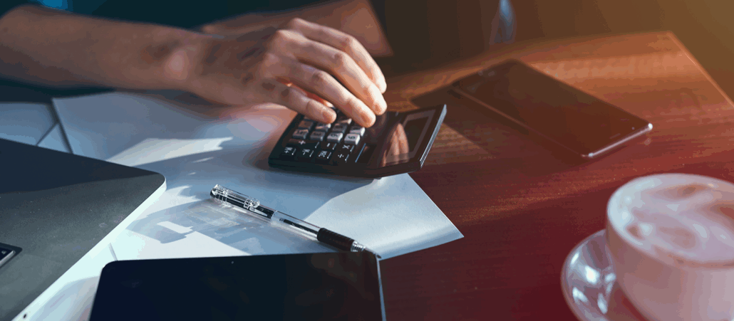 A close-up of a person’s hands using a calculator on a wooden desk. Papers, a pen, a laptop corner, a smartphone, and a cup of coffee surround the workspace, suggesting financial or accounting work.