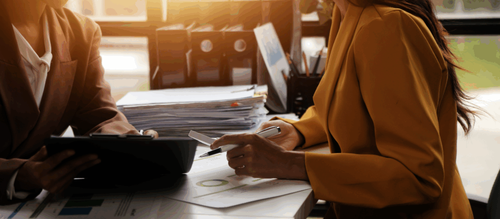Two professionals sitting at a desk reviewing paperwork and charts, with one holding a tablet and the other a pen, in a sunlit office environment.