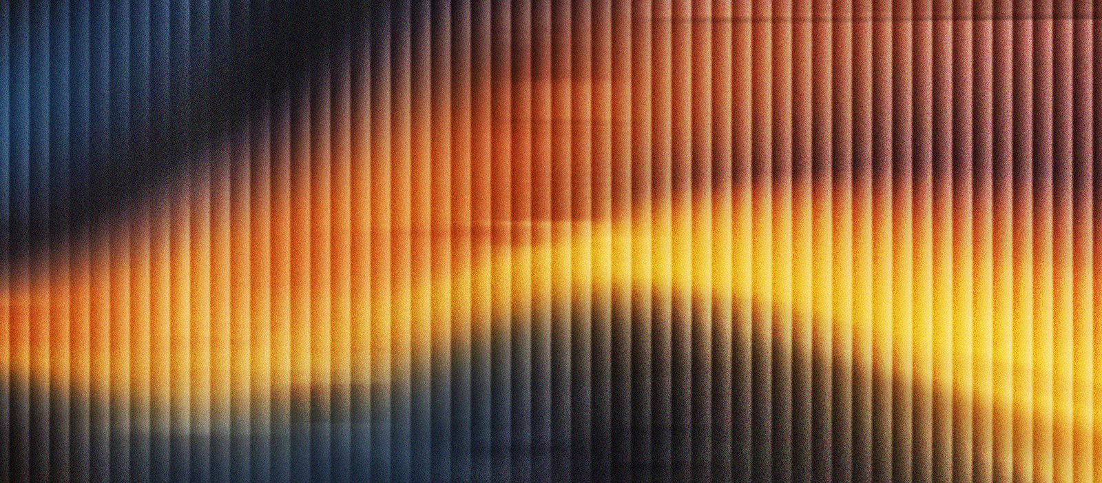 Abstract image of warm yellow and orange light blending into deep blue tones behind vertical ribbed glass, creating a soft, blurred gradient effect. The vertical lines add texture and a sense of depth across the composition.