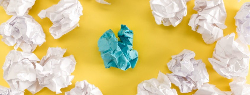 Alt text: Crumpled paper balls on yellow background with one blue paper in the center.