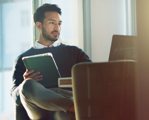A man sits in a modern office holding a tablet and looking at a laptop, with sunlight streaming through large windows and a small plant on a side table.
