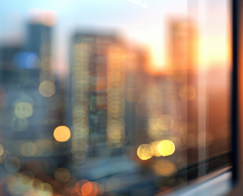 Blurred view of a city skyline at sunset seen through a glass window, with colorful lights and reflections out of focus.