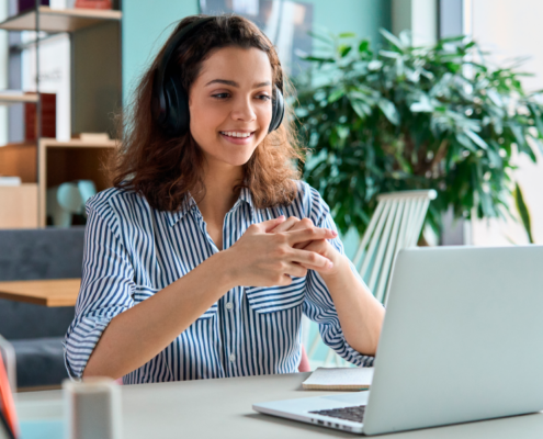 A woman wearing headphones smiles while working on a laptop in a bright, modern home office.