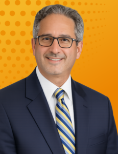 Portrait of a senior legal leader (Luis Machado) wearing glasses, a navy suit, and a striped tie against an orange background.