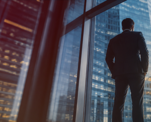 Silhouette of a businessman in a suit standing at a window, looking out over a city skyline of high-rise buildings at sunset.
