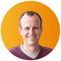 Headshot of Mark Kahn, a smiling man with short brown hair wearing a maroon crew-neck sweater, set against an orange circular backg